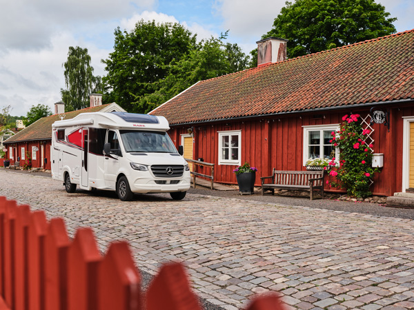 En KABE-husbil parkerad på en kullerstensgata framför charmiga röda trähus med blomsterdekorationer. En perfekt kombination av husbilsfrihet och svensk idyll.