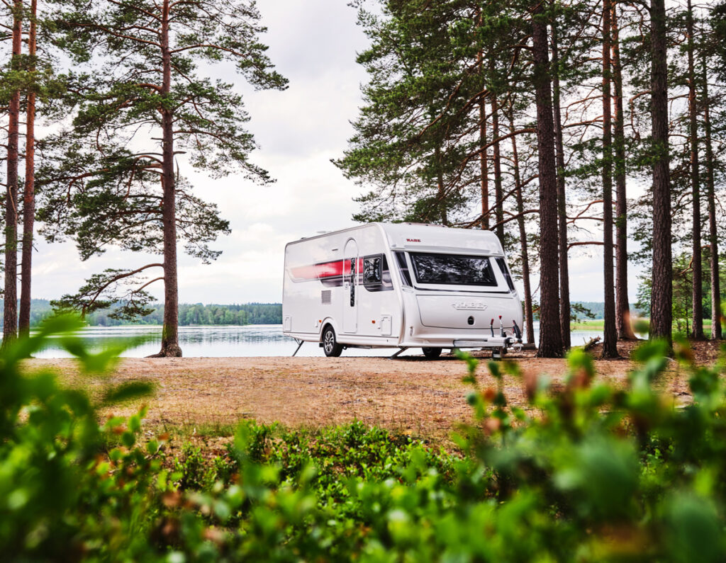 En KABE-husvagn står uppställd på en naturskön plats vid en sjö, omgiven av höga tallar. Den rymliga husvagnen smälter in i den lugna miljön, perfekt för avkoppling och camping nära naturen.