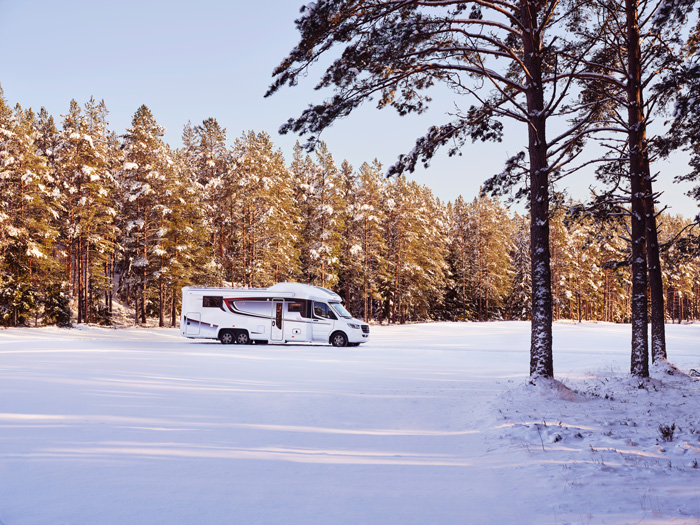 En KABE-husbil står parkerad i ett snötäckt landskap med solbelysta träd i bakgrunden. Perfekt för vintercamping i natursköna miljöer.