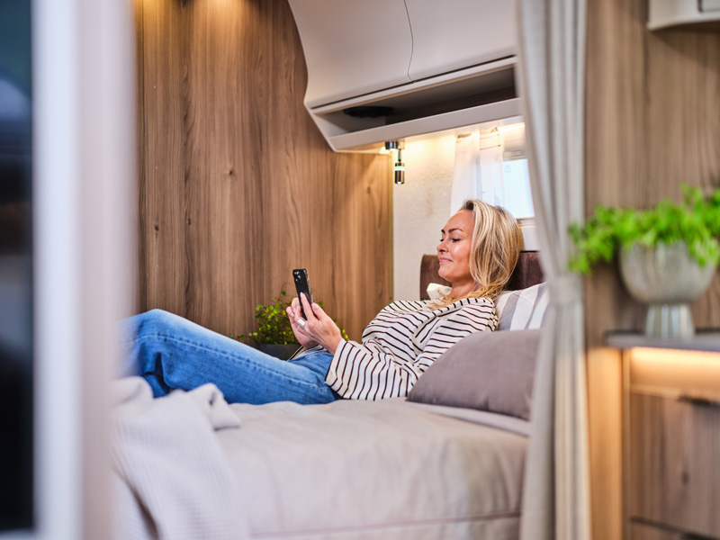 En kvinna ligger avslappnat på en säng i en husbil eller husvagn och tittar på sin mobil. Interiören har en elegant trädesign, mjuk belysning och dekorativa växter, vilket skapar en mysig och hemtrevlig atmosfär.