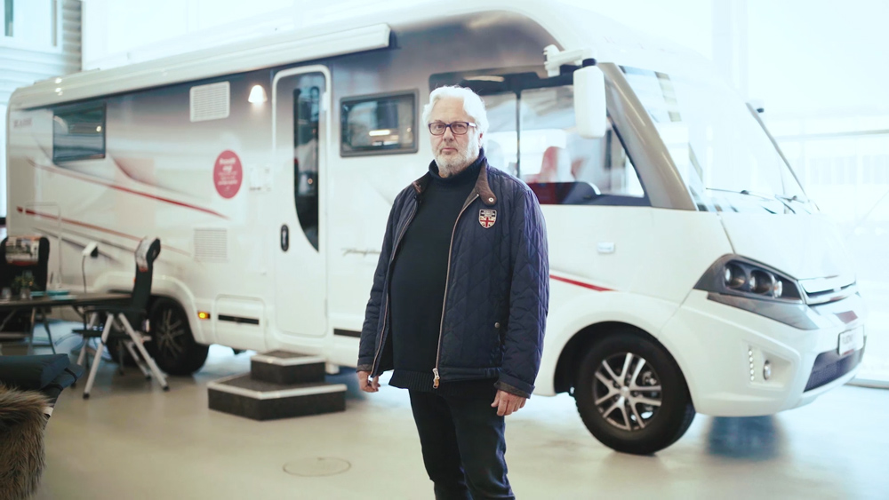 Peter Rudvi står inomhus framför en KABE-husbil i en ljus utställningshall. Han bär en mörkblå quiltad jacka med ett brittiskt emblem och tittar mot kameran. I bakgrunden syns campingmöbler och ett uppställt trappsteg vid husbilens entré.