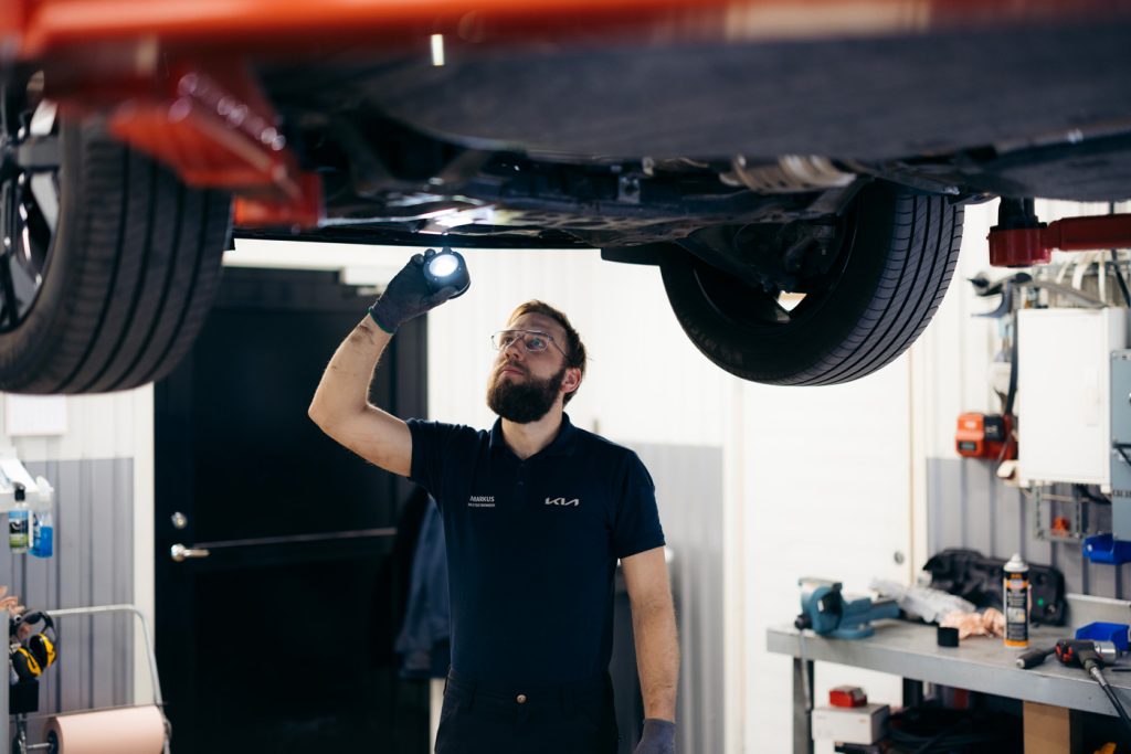 Kia-tekniker inspekterar undersidan av ett fordon med ficklampa i en verkstadsmiljö.