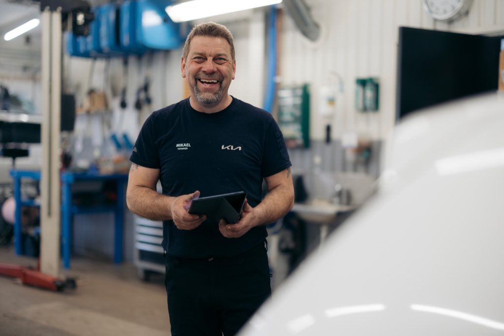 Mekaniker i verkstadsmiljö ler mot kameran med en surfplatta i handen – ett exempel på personlig service och teknisk expertis hos Kia-återförsäljaren Rudvi Bil i Blekinge.