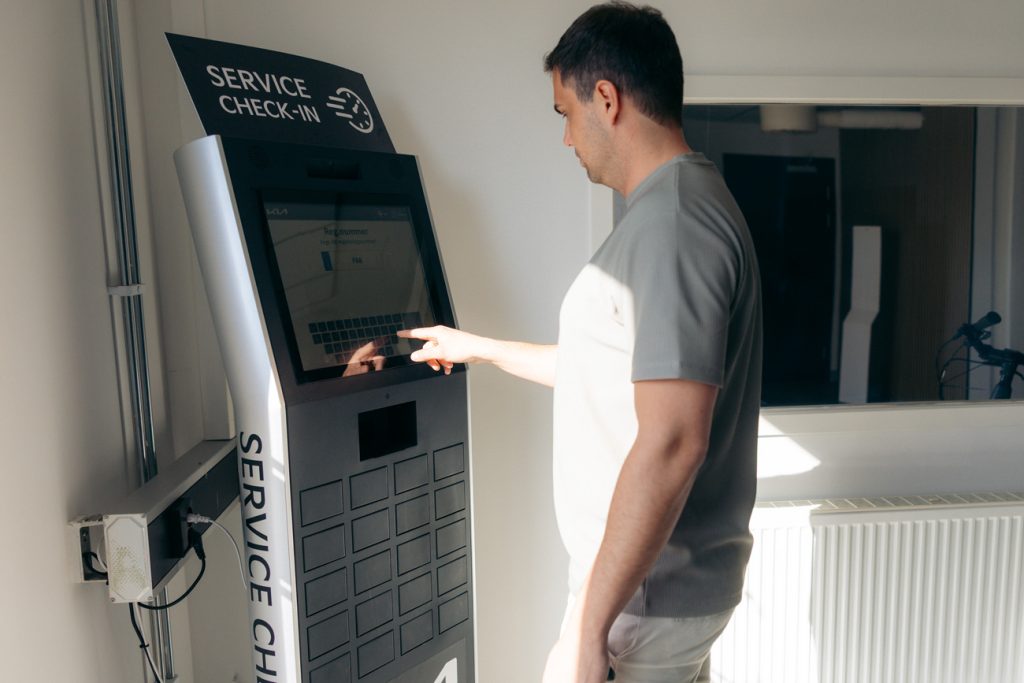 Man står vid Tjekviks självbetjäningskiosk hos Rudvi Bil och trycker på skärmen för att checka in bilen till service. Kiosken är placerad vid serviceingången och används för digital in- och utcheckning.