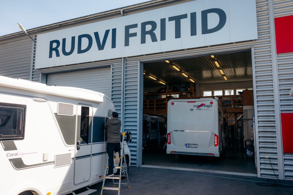 En person står på stege och utför arbete på en husbil utanför verkstadsporten till Rudvi Fritid. I bakgrunden syns en annan husbil inne i verkstaden under företagets skylt.