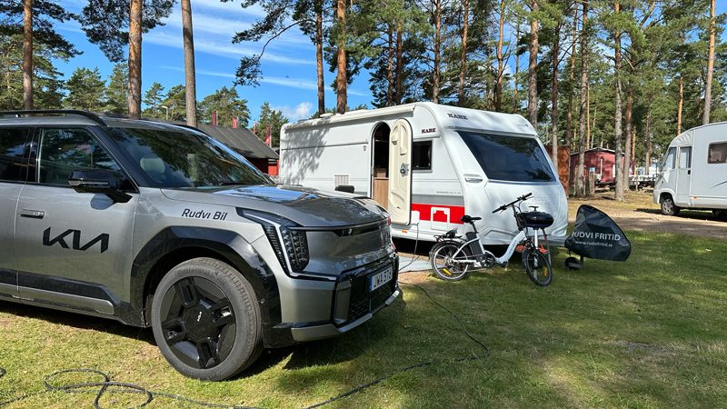 Elbil Kia EV9 laddar elcykel vid husvagn på camping – exempel på att dra husvagn med elbil och använda bilen som strömkälla för eldrivna fordon.
