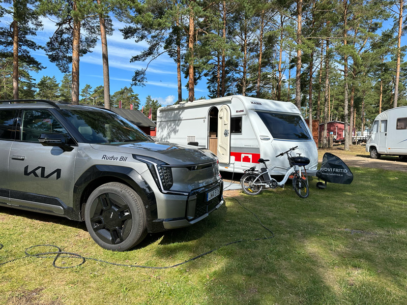 Elbil Kia EV9 laddar elcykel vid husvagn på camping – exempel på att dra husvagn med elbil och använda bilen som strömkälla för eldrivna fordon.